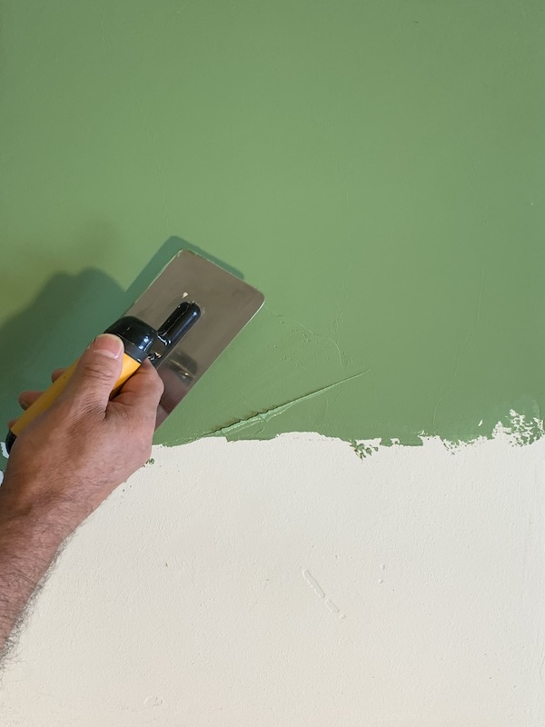 Eine Hand trägt mit einer angewinkelten Edelstahl-Glättekelle die erste dünne Schicht Stucco Veneziano auf eine glatte Wand auf